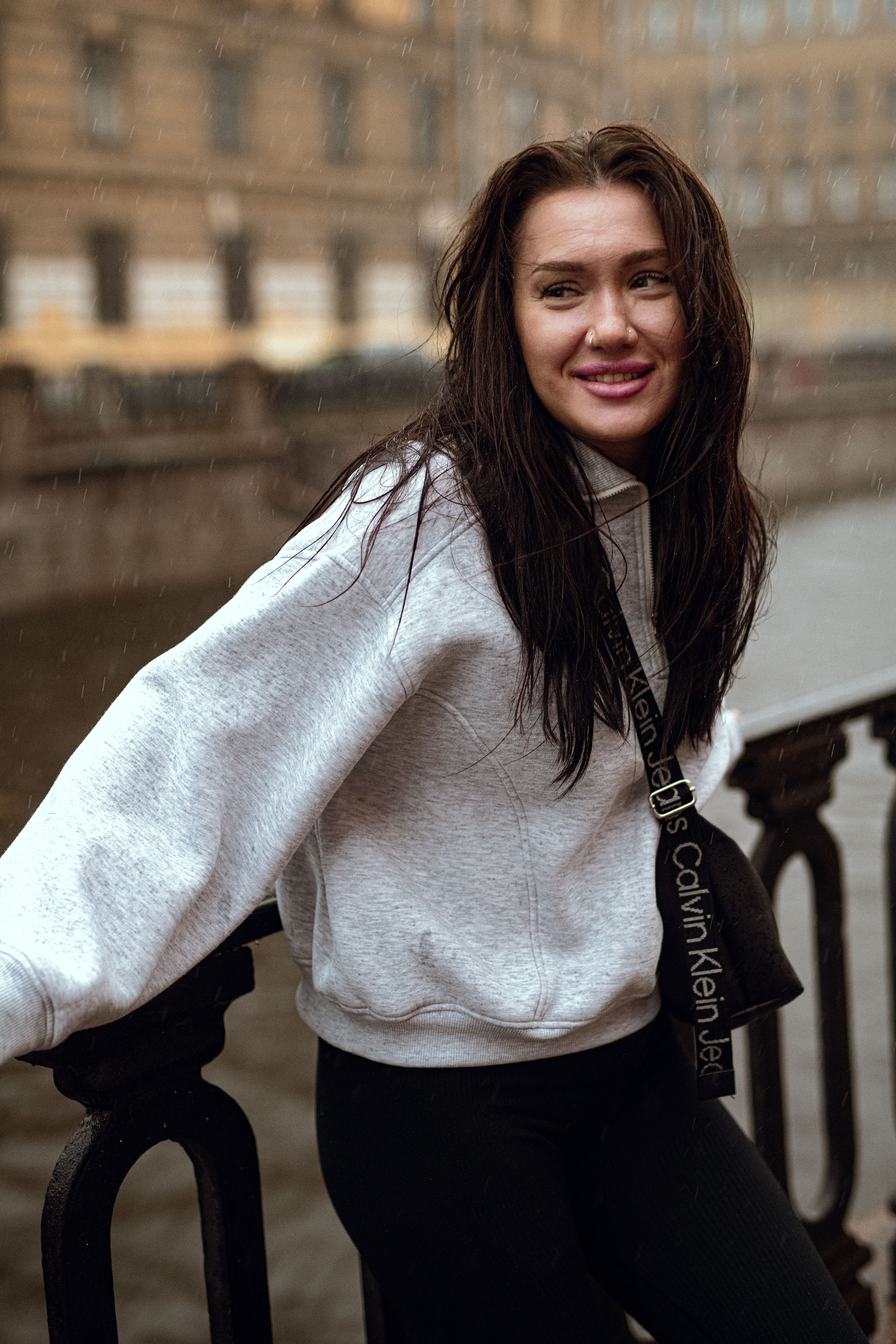 Smiling woman in a white hoodie leaning against a railing in a rainy cityscape.