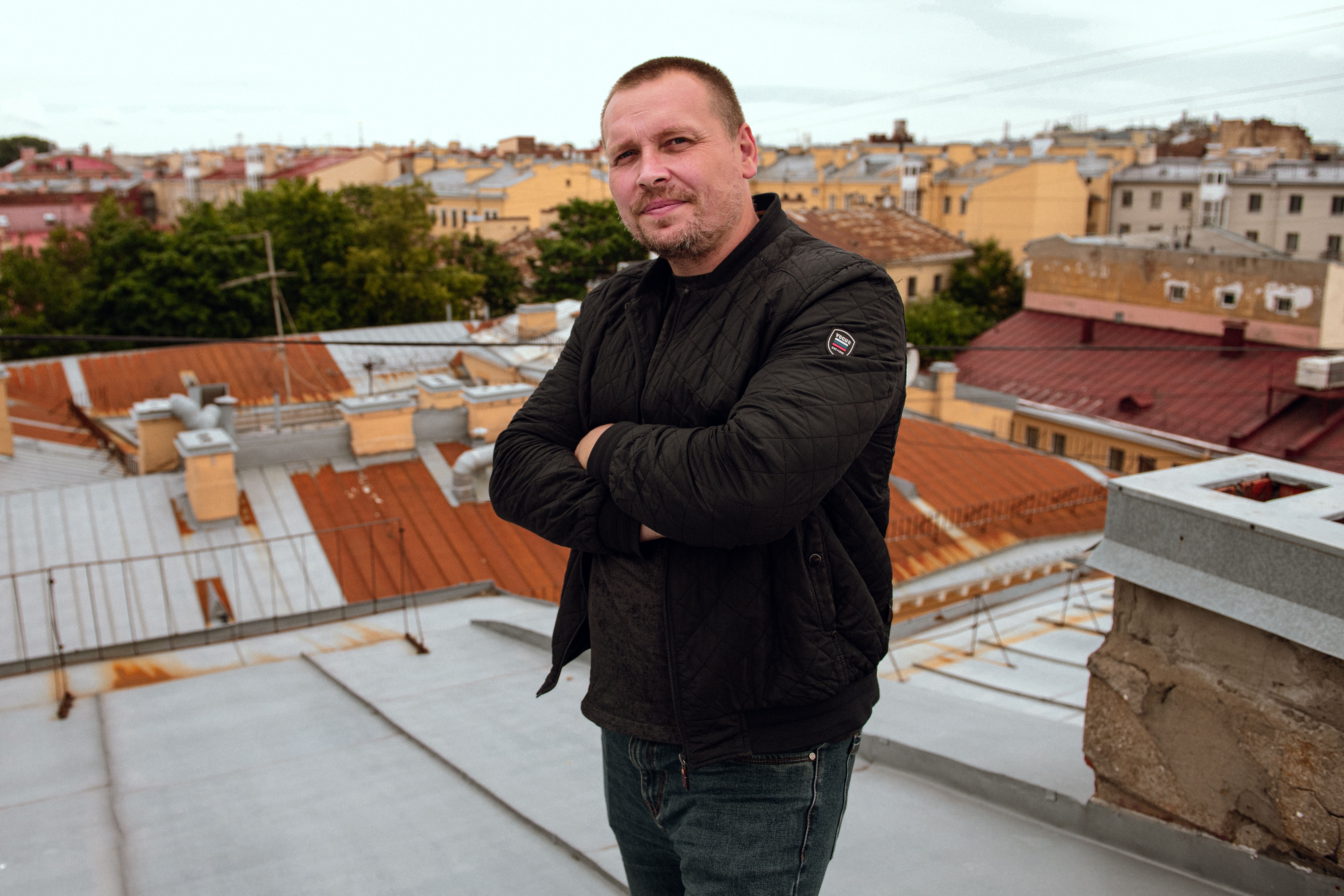 Man in black jacket confidently stands on rooftop with cityscape backdrop on a cloudy day.