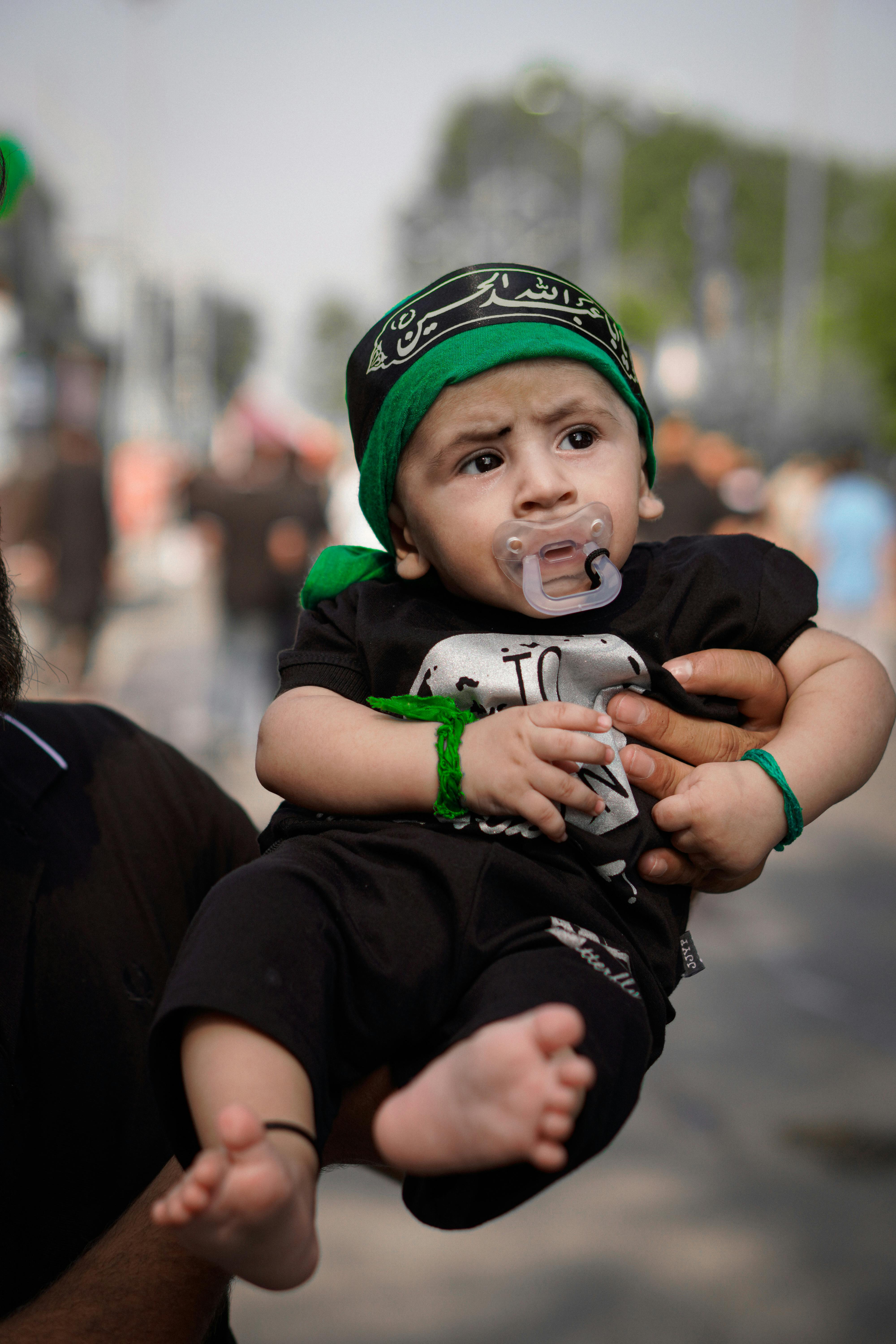 Free Cute baby boy wearing a green headband and black outfit being held outdoors at a festival. Stock Photo