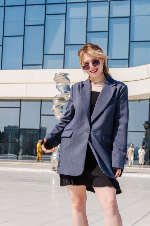 Free Stylish woman in sunglasses and blazer poses outdoors against a sleek glass building facade. Stock Photo