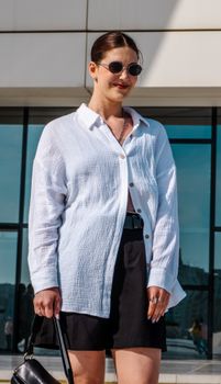 Fashionable woman in sunglasses wearing a chic white shirt and black shorts outdoors.
