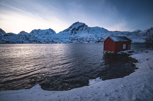 Free Tranquil winter scene featuring a red cabin by a lake with snowy mountains at dusk. Stock Photo