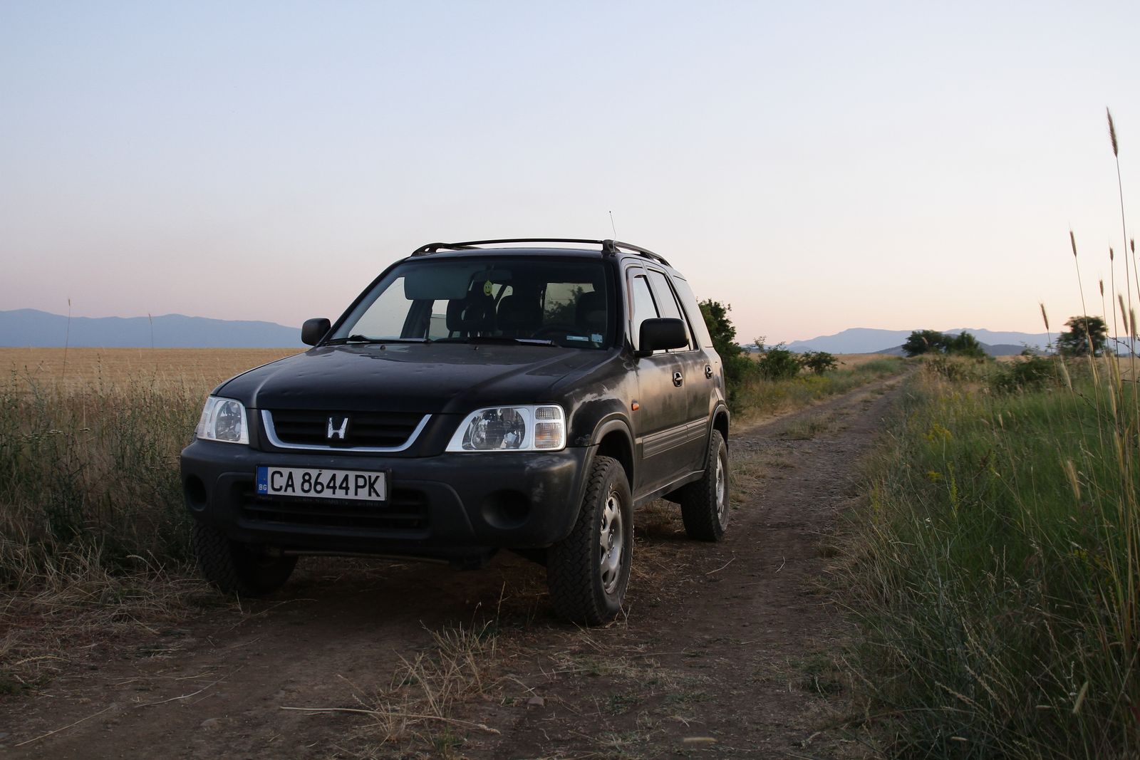 Black SUV on a dirt road surrounded by fields at sunset, capturing a serene rural landscape.