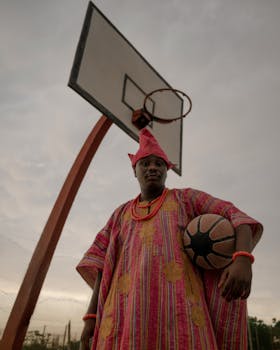 Man in traditional Nigerian attire holding a basketball under a hoop.