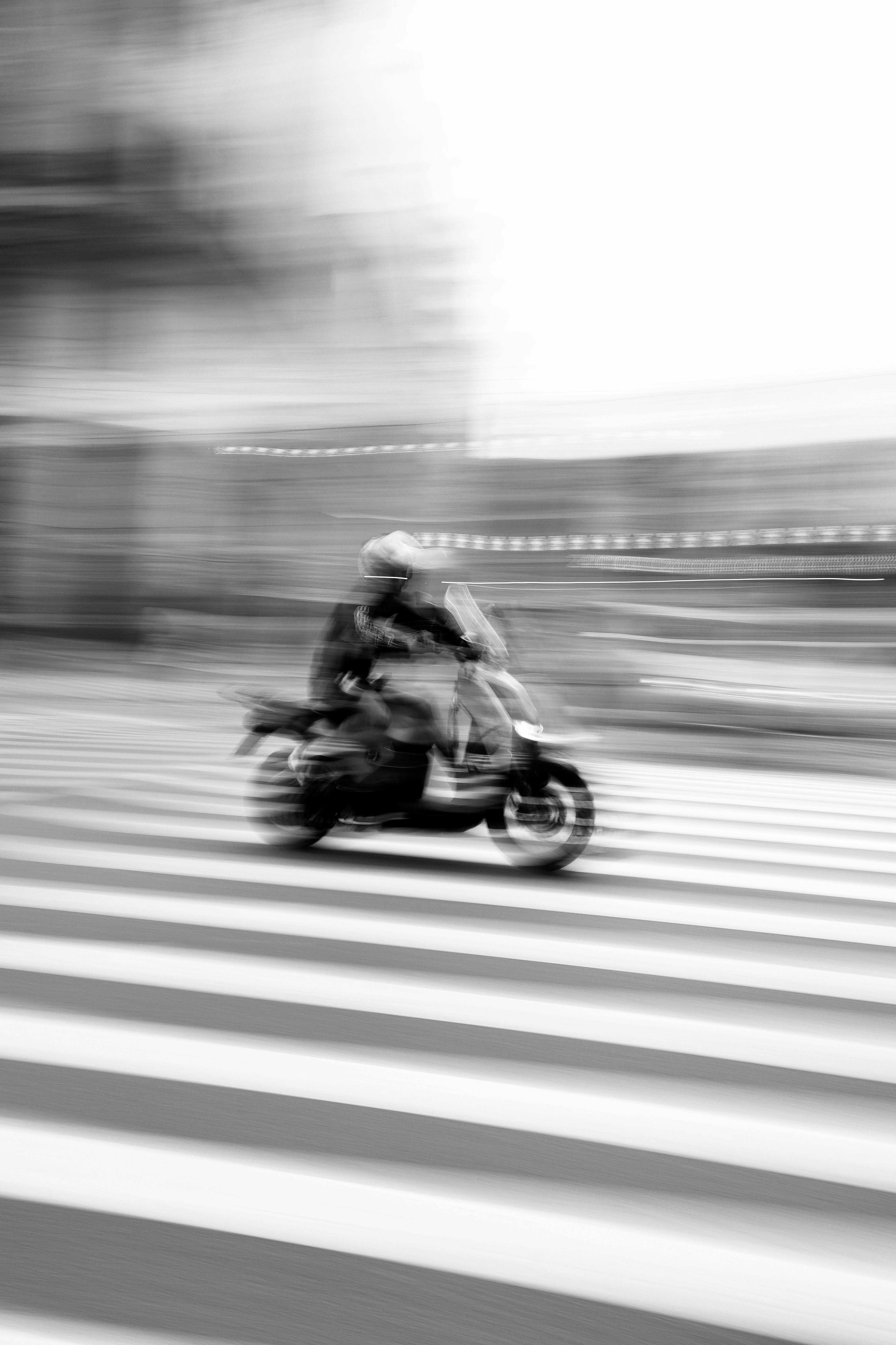 Black and white photo capturing blurred motorcycle crossing a street, illustrating urban motion.