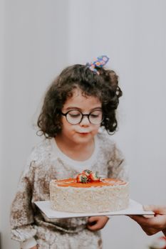 Adorable child with glasses blows out a birthday cake candle indoors.