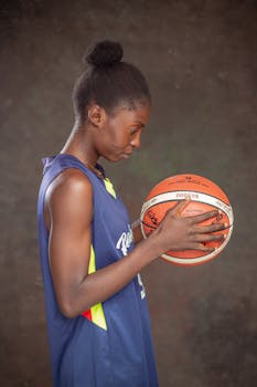A young female basketball player in a blue jersey focused on a basketball, with a thoughtful expression.