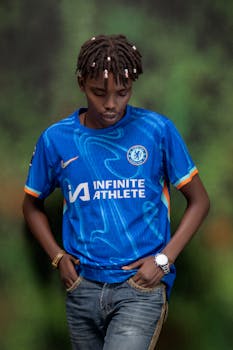 A young man poses confidently in a blue Chelsea FC football jersey with hands in pockets against a blurred natural backdrop.