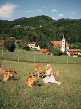 A woman sits among deer in a picturesque Slovenian landscape with hills and a village church.