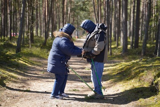 Elderly couple in winter coats enjoying a forest walk with trekking poles in Latvia.