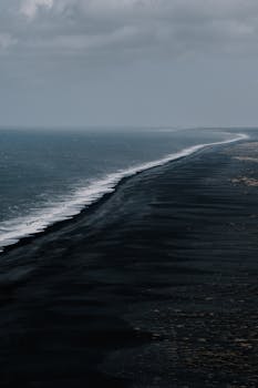 A dramatic aerial photograph of a black sand beach stretching along a stormy coastal line.