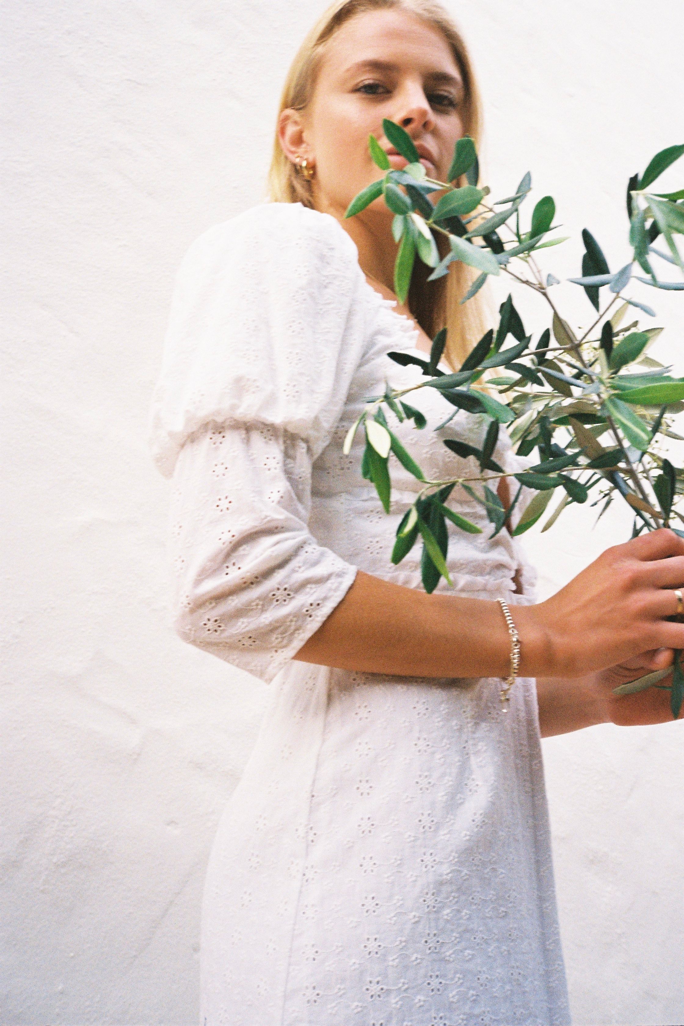 A woman in a white dress holding green leaves, standing against a white backdrop.