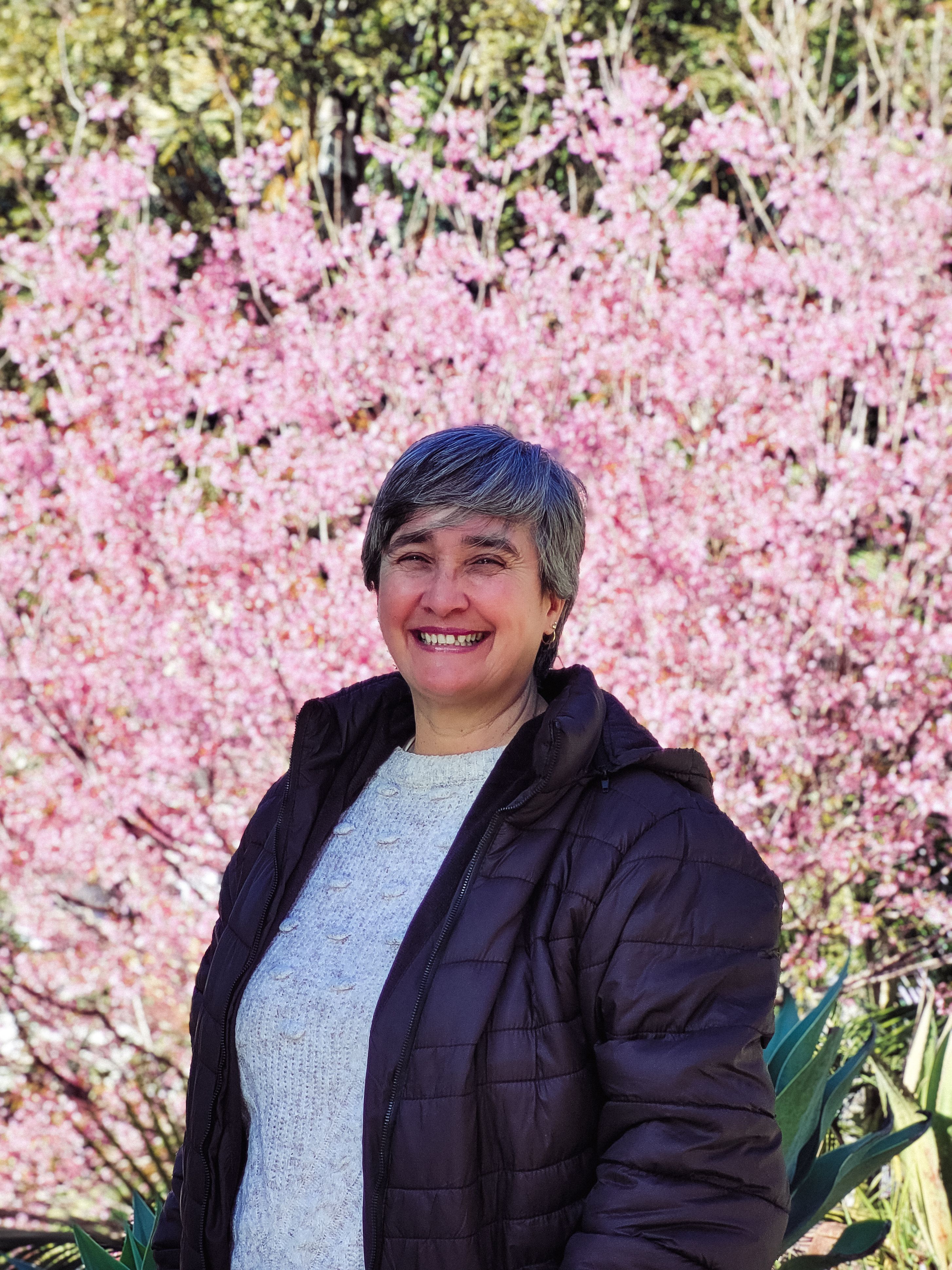 Happy woman in winter coat stands before vibrant cherry blossoms in a garden setting.