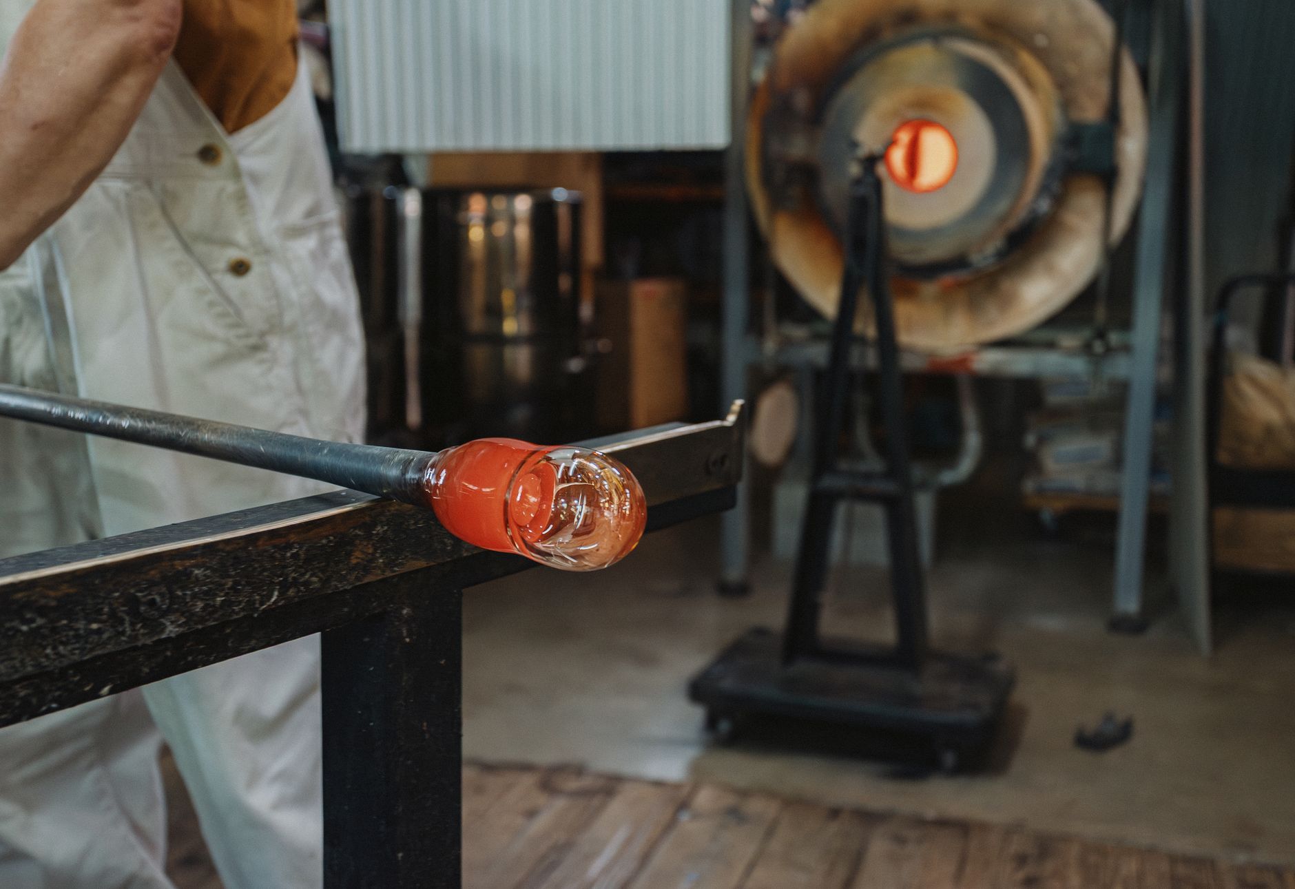 A maker steadying a piece of molten glass on the iron at the bench, the glory hole glowing behind
