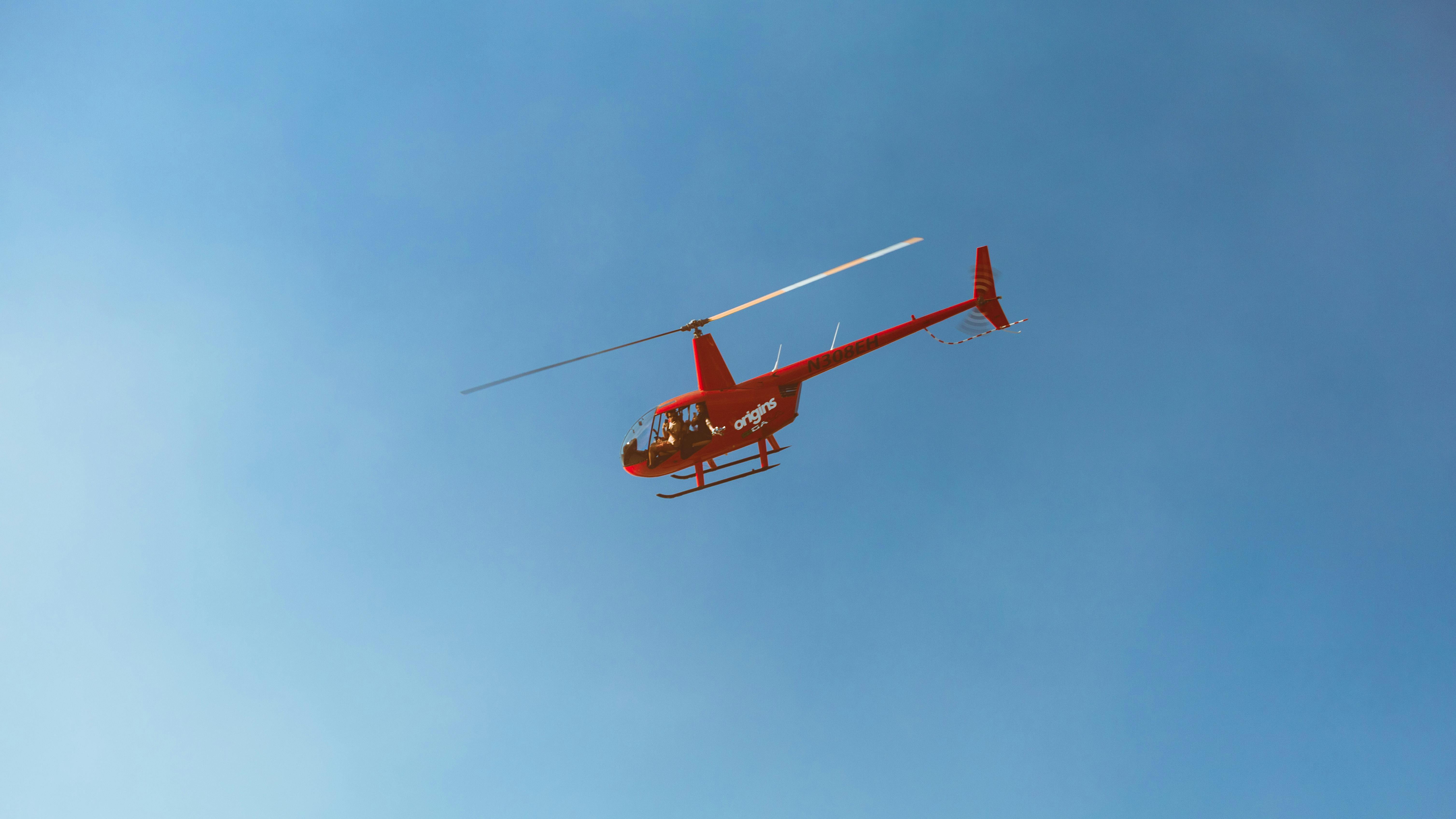 A red helicopter with 'origins' branding flying against a clear blue sky.