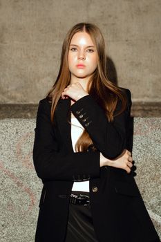 Chic young woman in a black blazer stands confidently against a textured wall in natural light.