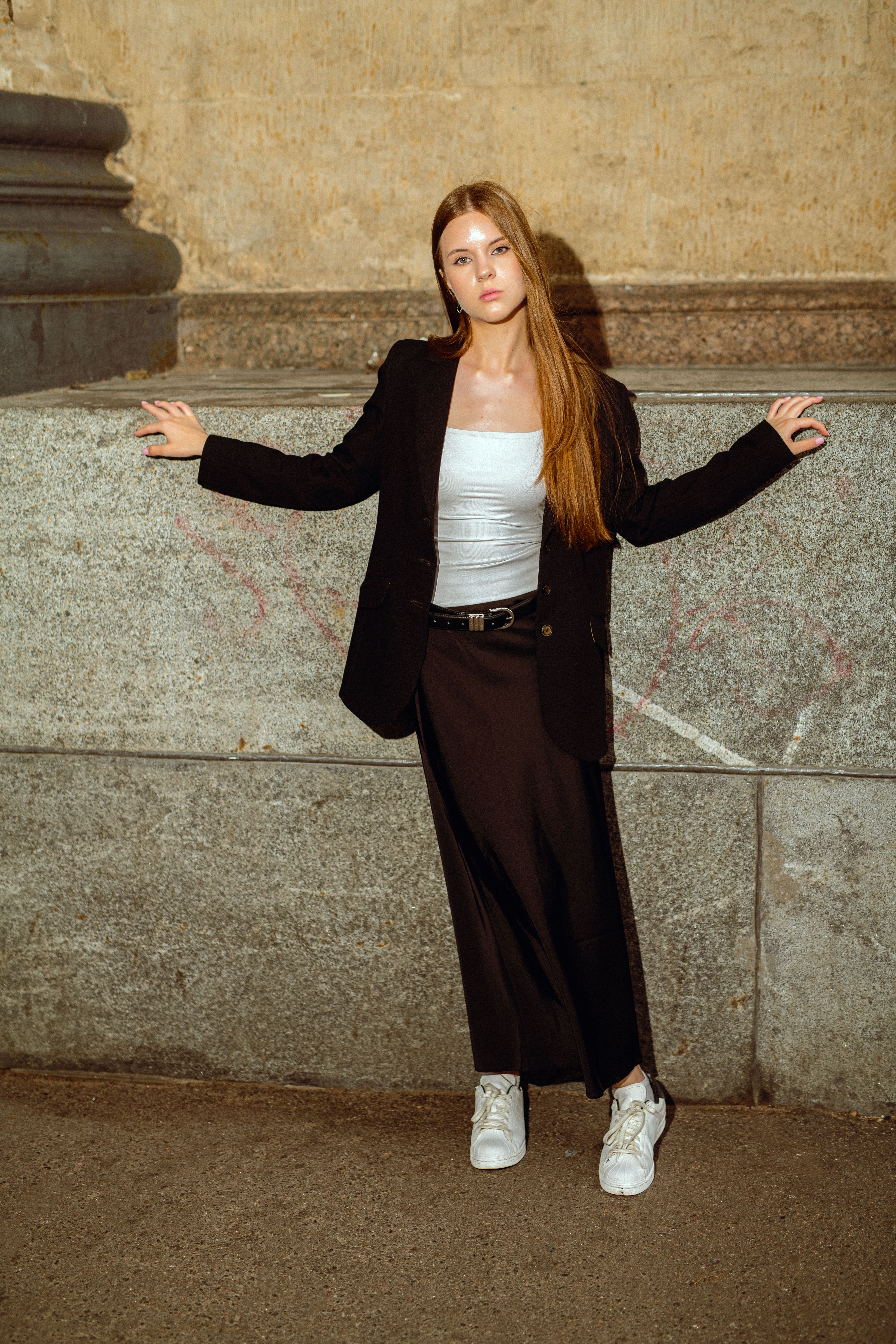 Fashionable woman in black blazer and skirt standing against a textured stone wall, exuding confidence and style.
