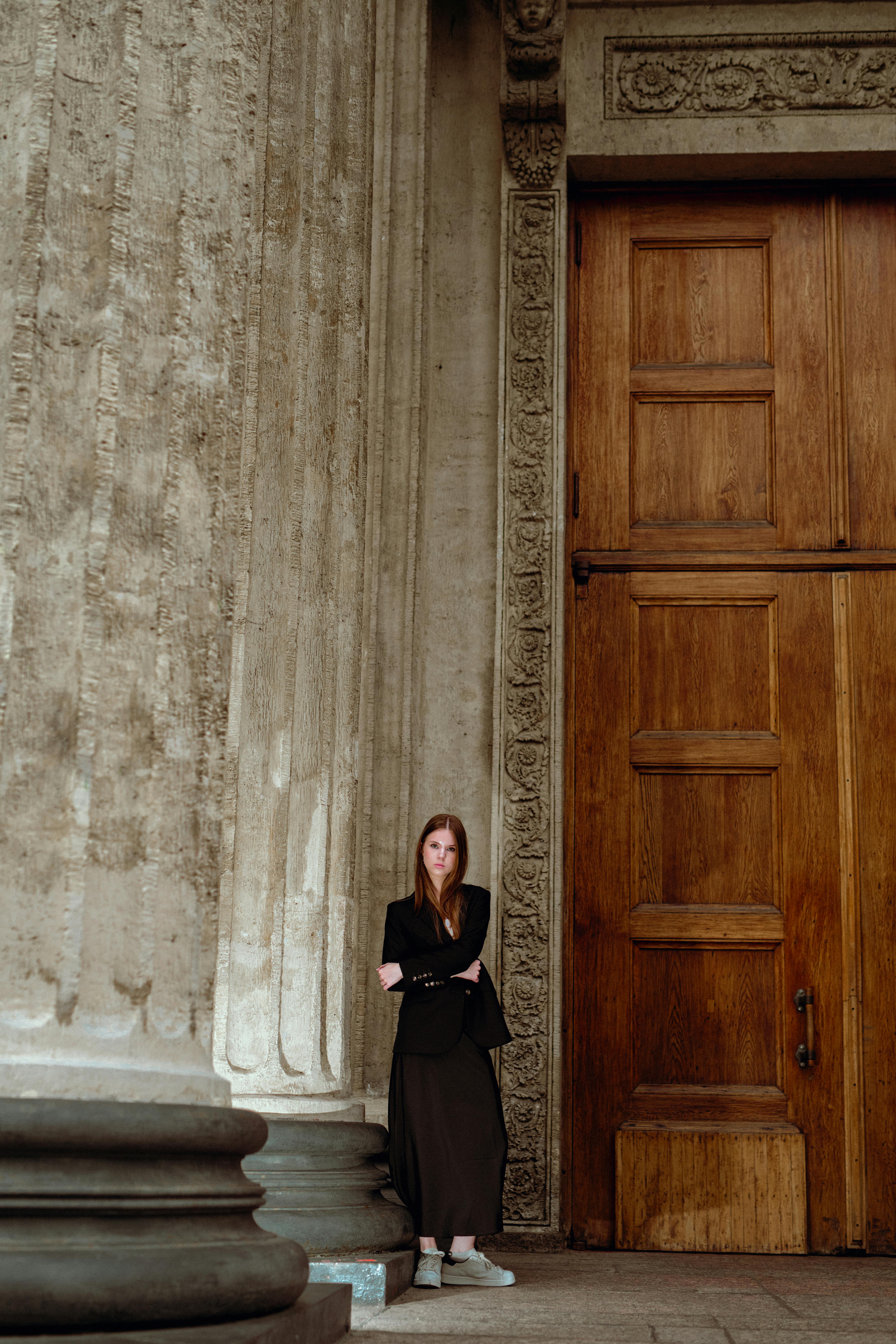 Free A woman in fashionable attire leans against a large wooden door next to detailed stone pillars. Stock Photo