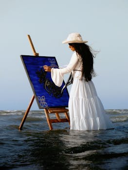 A young woman artist painting on the beach, standing in water, wearing a long white dress and hat.