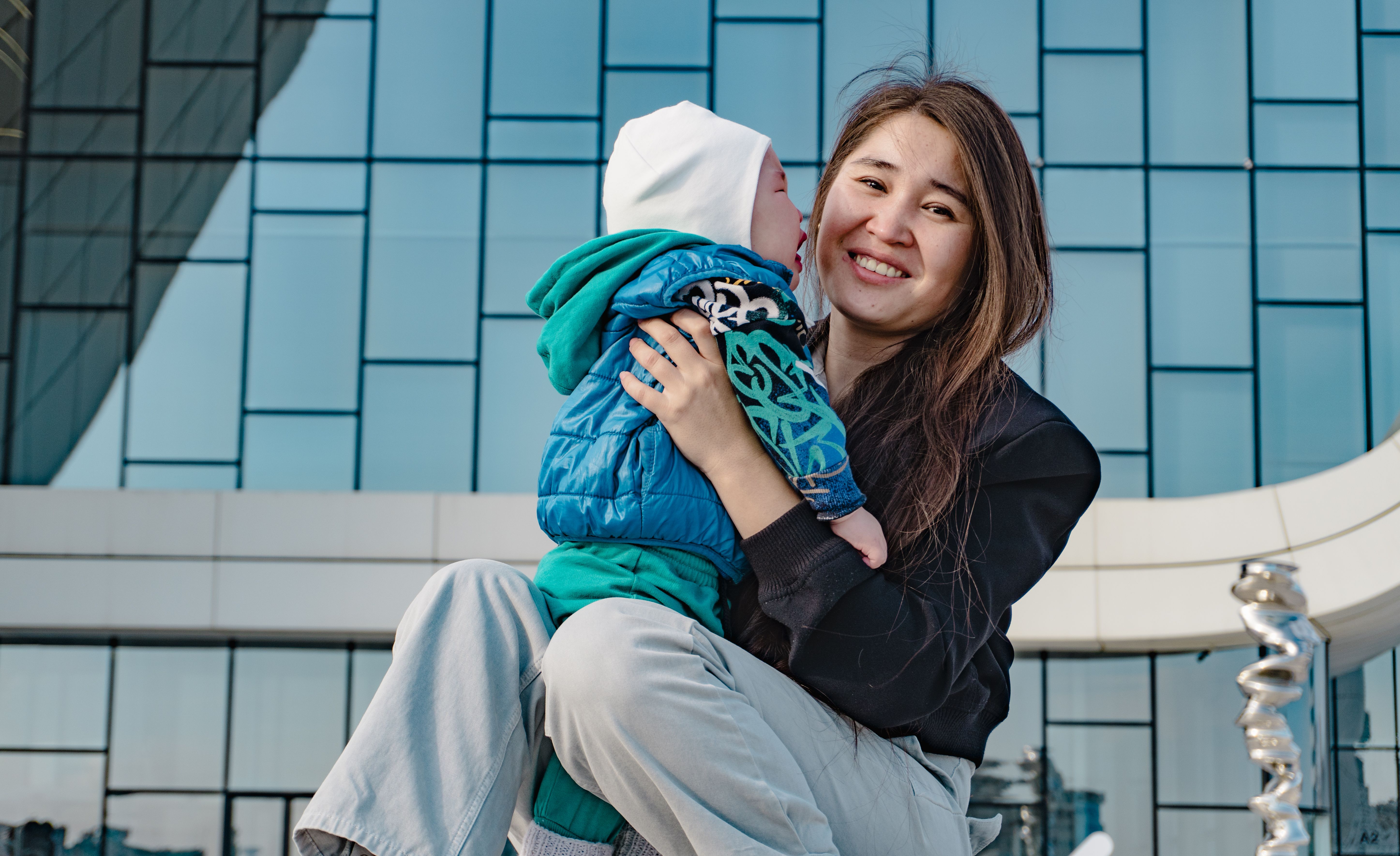 A joyful mother holding her baby outside a modern glass building, capturing a happy moment.