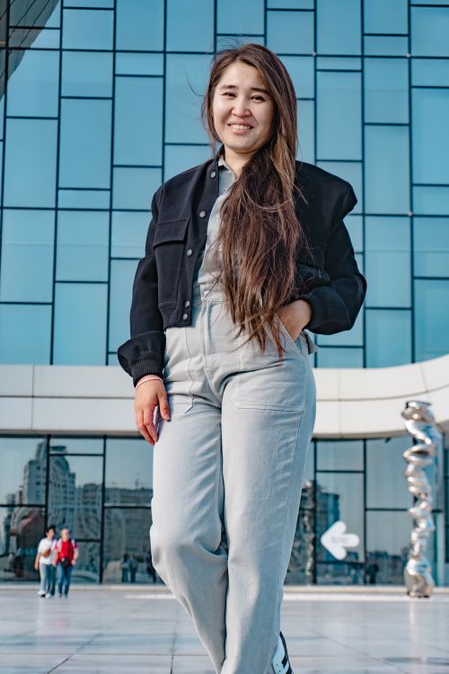 Free Young smiling woman posing confidently in front of a modern glass building, showing urban lifestyle. Stock Photo