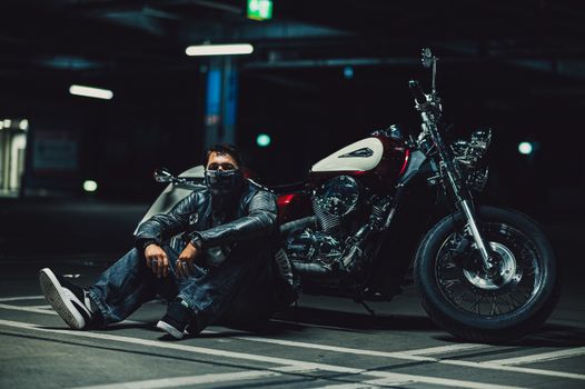 A biker wearing a leather jacket and mask sits beside a motorcycle in a dimly lit parking garage.