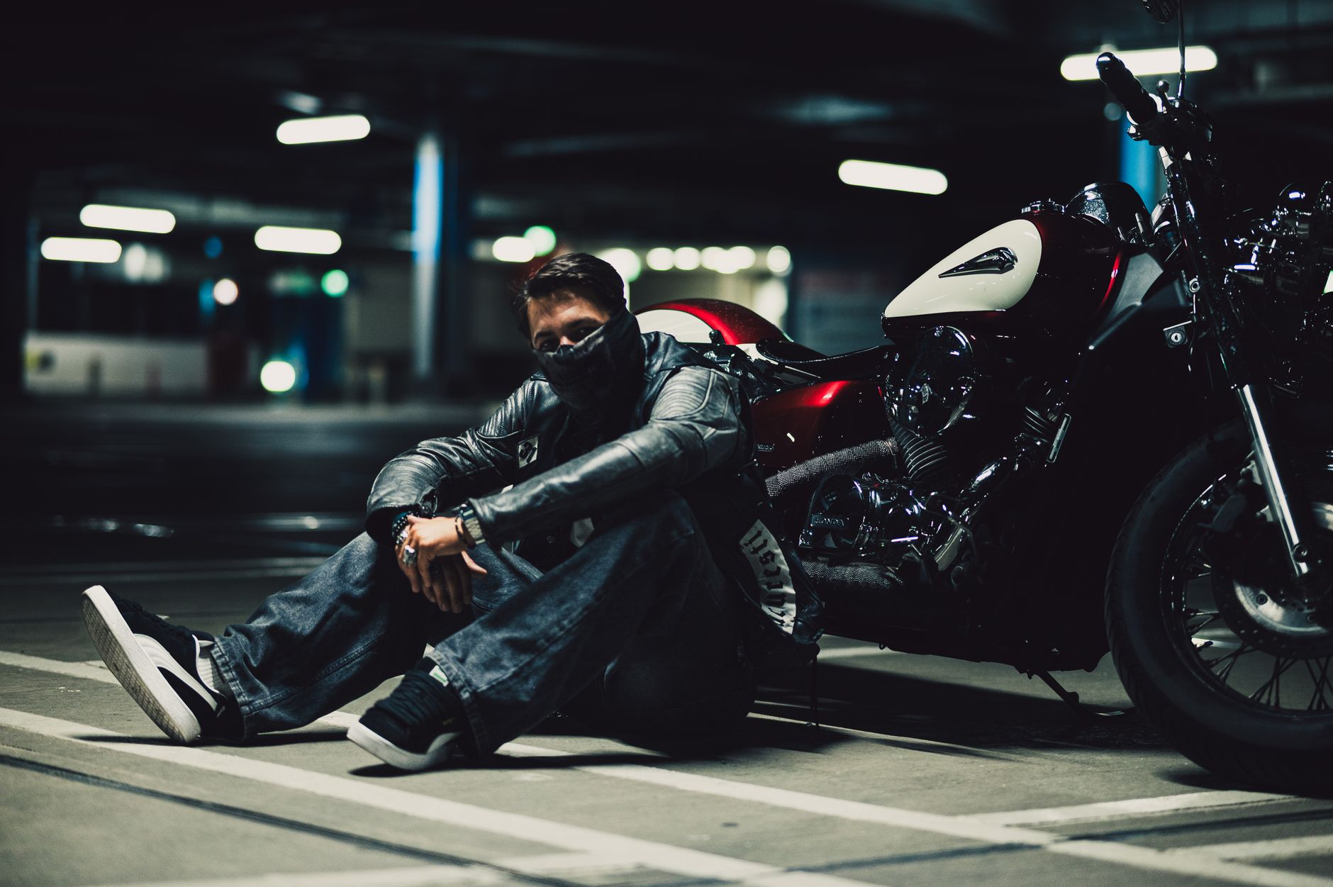A biker sits on the ground in a dimly lit parking garage beside a sleek motorcycle.