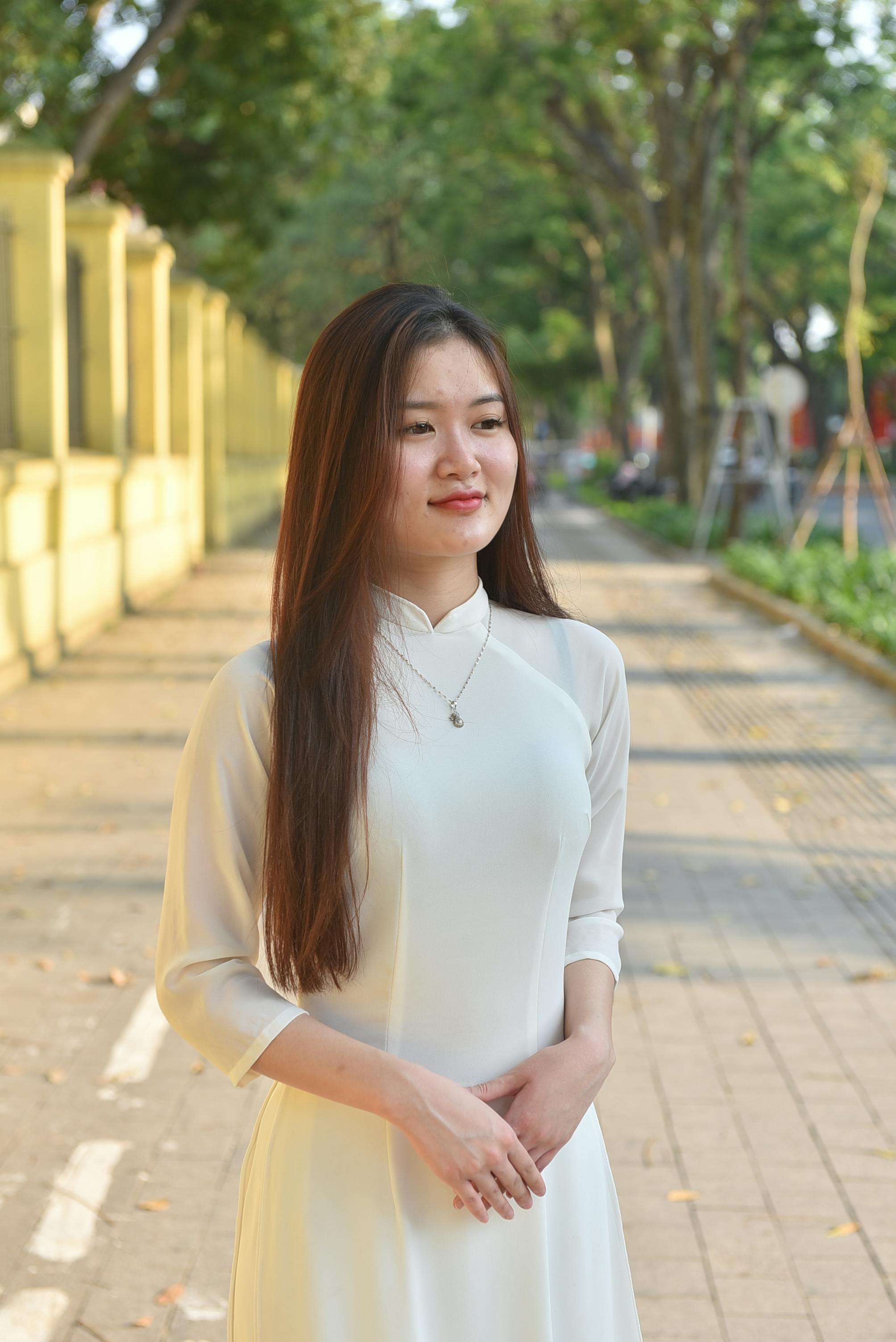 Elegant young woman in white Ao Dai dress standing on a sunny pathway.