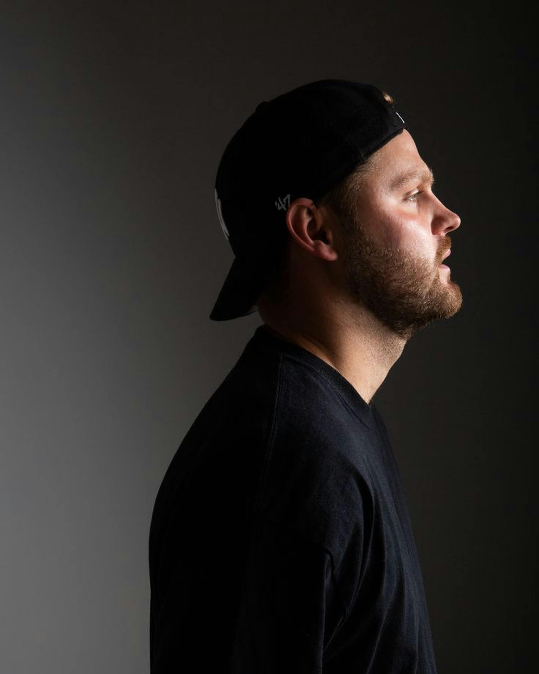 Side profile of a man in a black cap against a dark background.