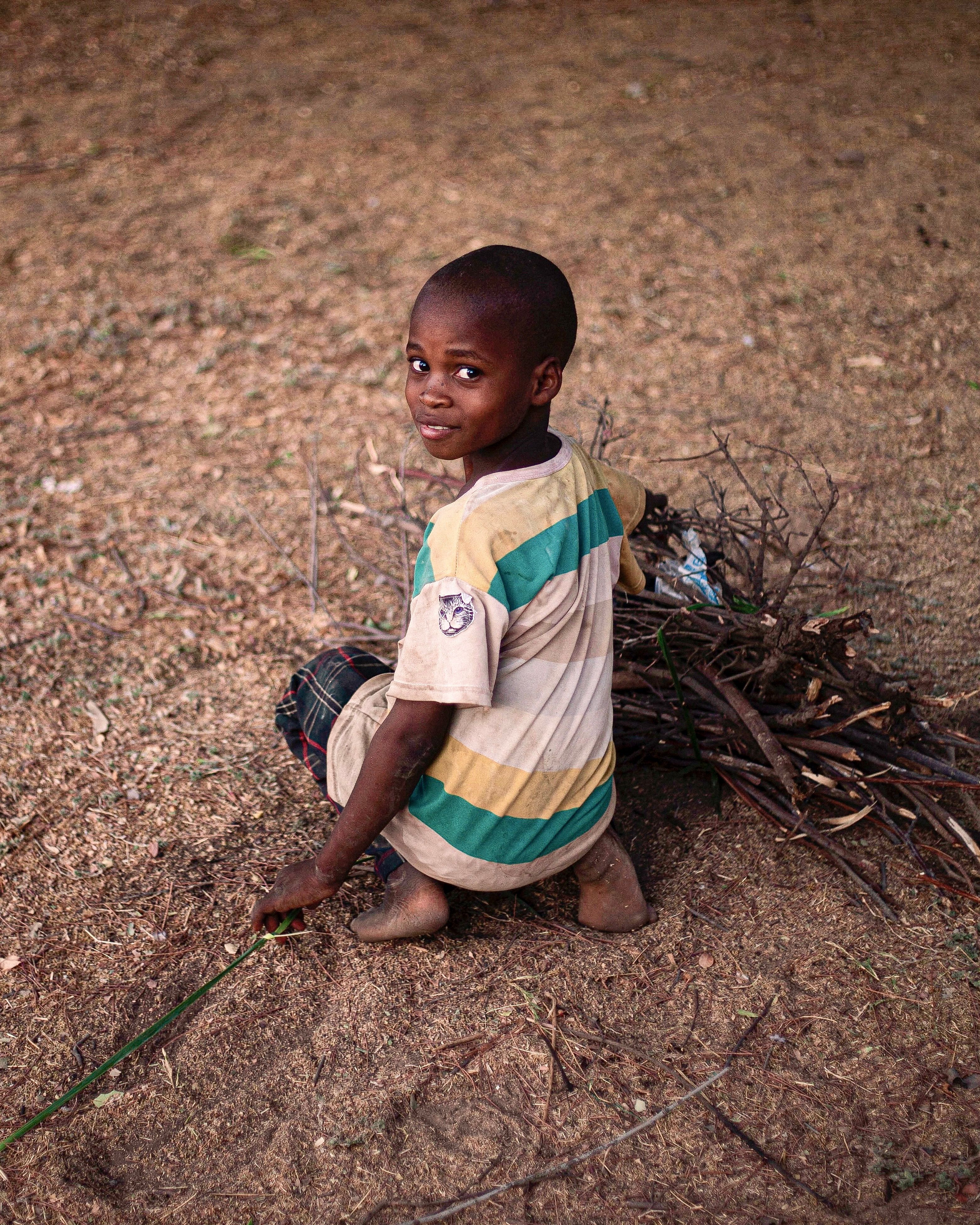 Young boy sitting outdoors collecting firewood, smiling at camera.