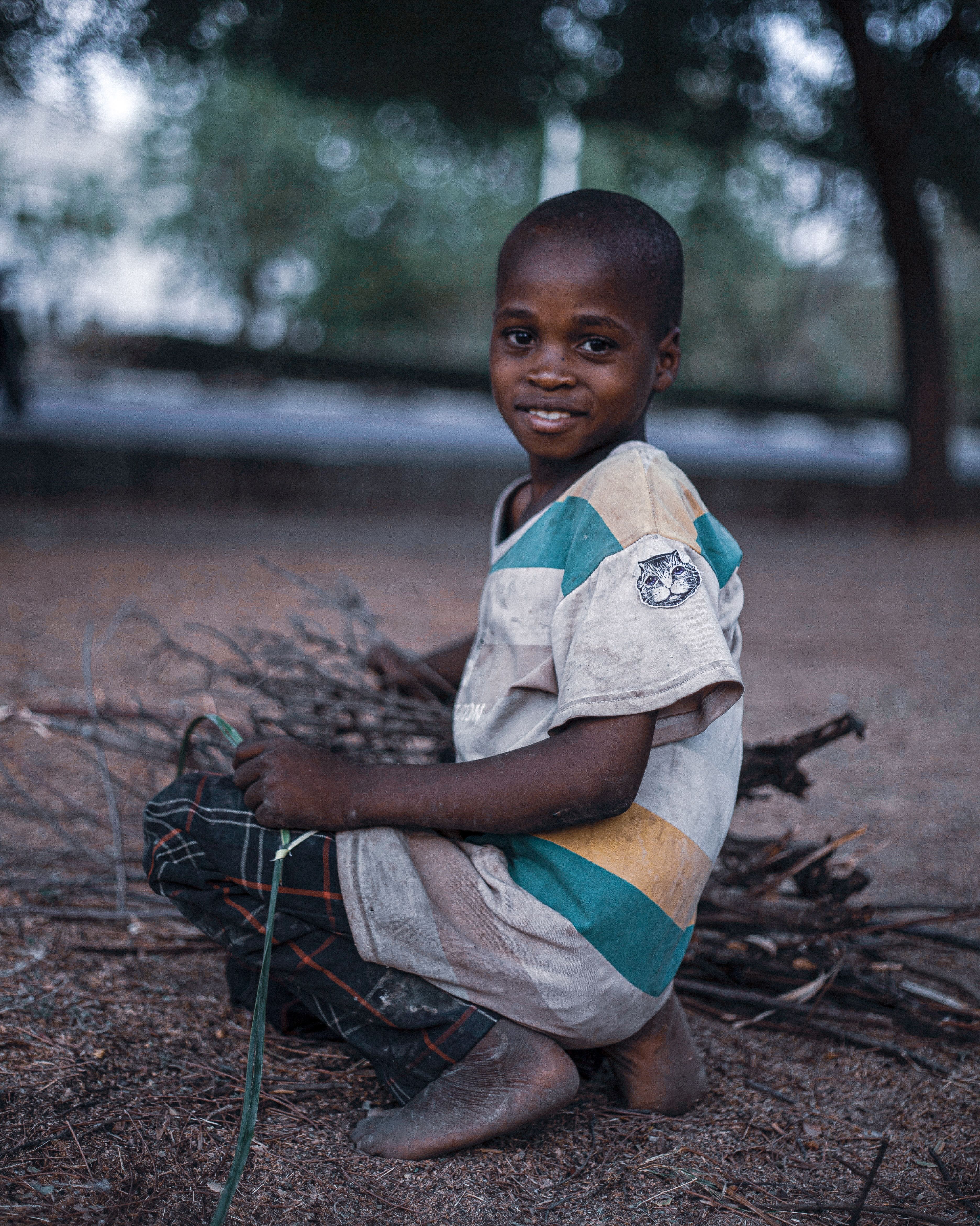 A young boy in striped shirt gathers firewood outdoors, smiling at the camera.