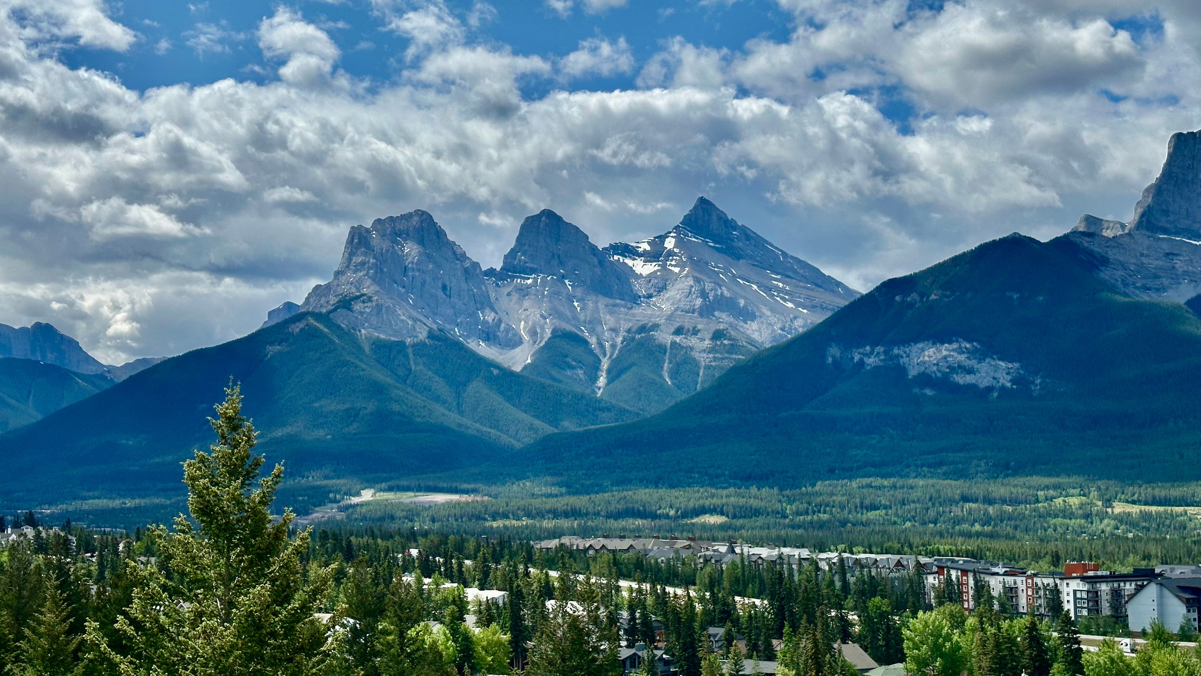 Three jagged peaks rise above a forested valley with buildings at its foot, under a cloudy blue sky