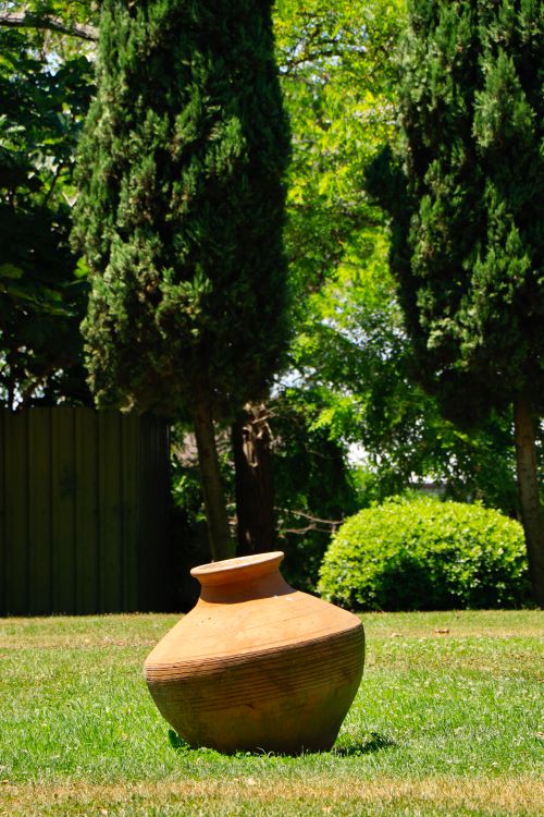 Free A rustic clay pot on grass surrounded by lush greenery and trees under bright sunlight. Stock Photo