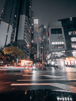 Dynamic city street at night with towering skyscrapers illuminated by city lights and reflections on wet pavement.