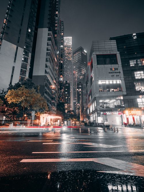 Free Dynamic city street at night with towering skyscrapers illuminated by city lights and reflections on wet pavement. Stock Photo