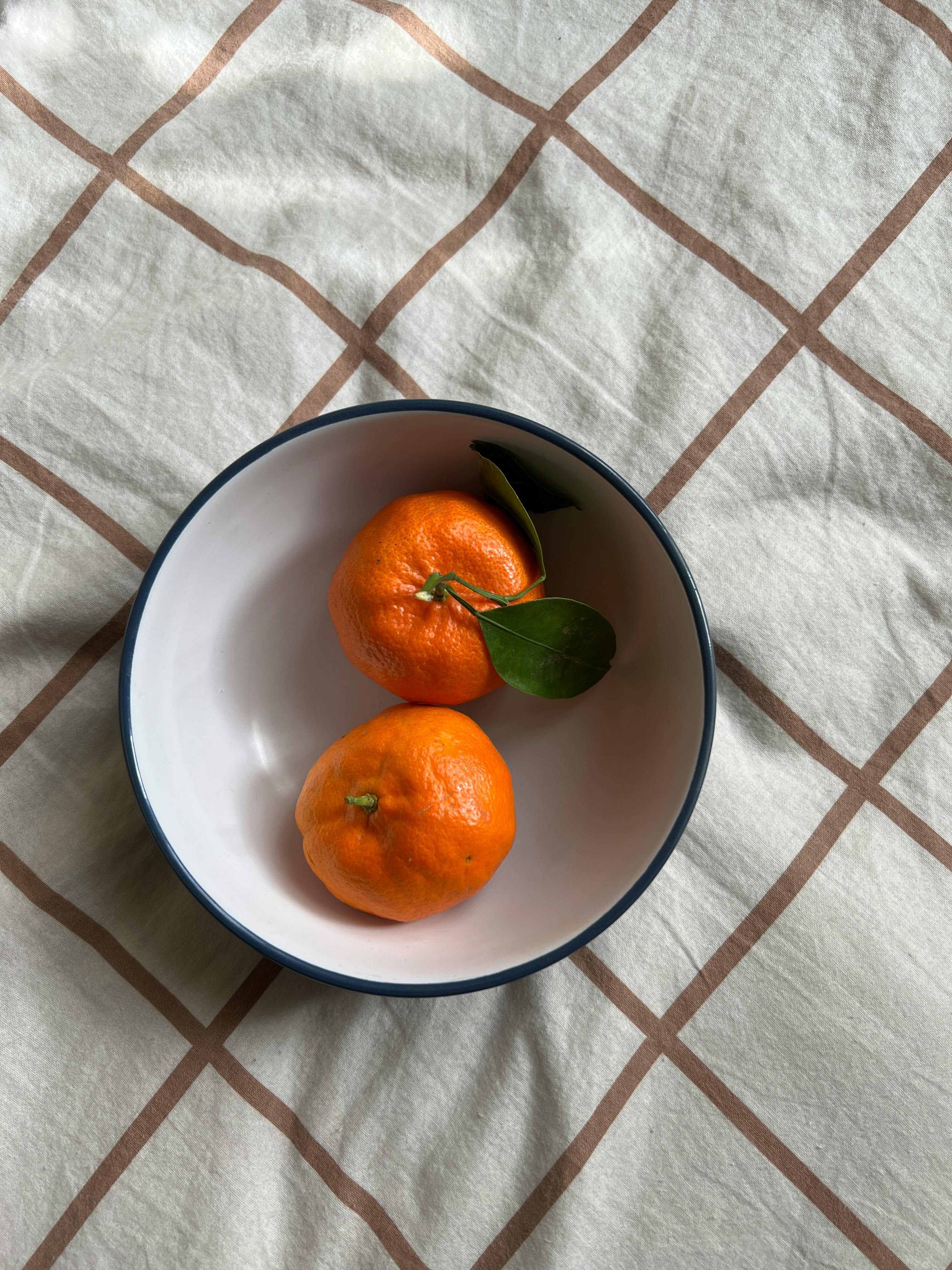 Two ripe tangerines with green leaves in a bowl on a checkered background, bright and fresh.