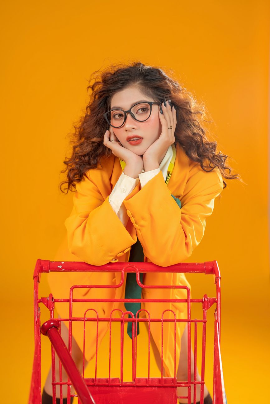 Stylish woman in bright outfit poses with shopping cart against vibrant orange background.