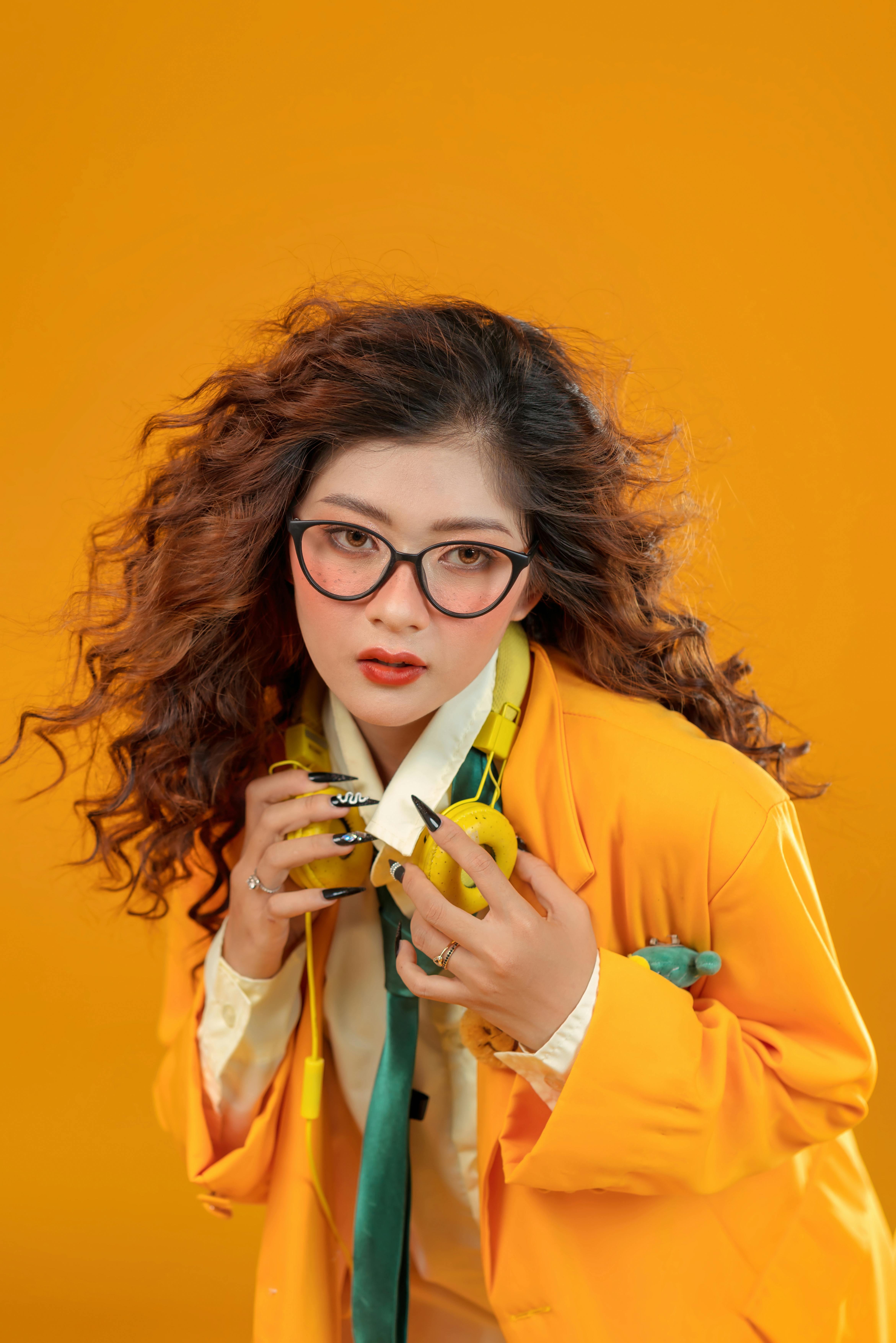 Kostenlos Modische Frau mit Brille, Anzug und Kopfhörern posiert vor einem kräftigen orangefarbenen Hintergrund. Stock-Foto