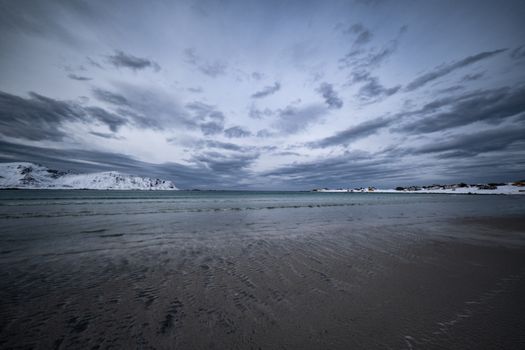 Moody beach scene showing snow-covered mountains under dramatic clouds.
