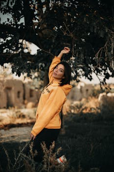 一名时尚女子在户外树下穿着黄色连帽衫摆姿势