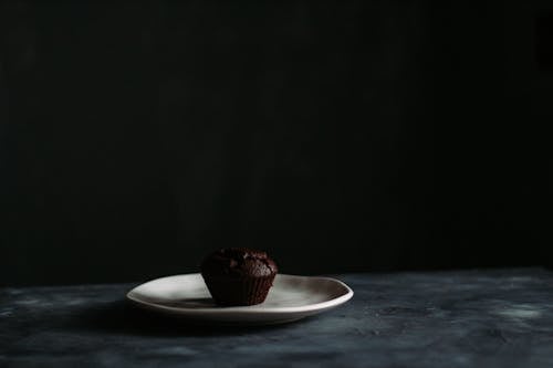 Chocolate muffin served on white ceramic plate placed on marble table against black background illuminated by daylight