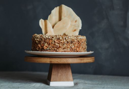 Yummy homemade honey cake decorated with nut mix and thin white chocolate tablet on top served on ceramic plate placed on wooden pedestal cake plate located on tablecloth against black background in daylight