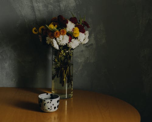 Various colors and kinds and sizes buds of flowers collected in lush bouquet put in tall transparent vase with water placed on wooden table with spotted cup against dark gray wall