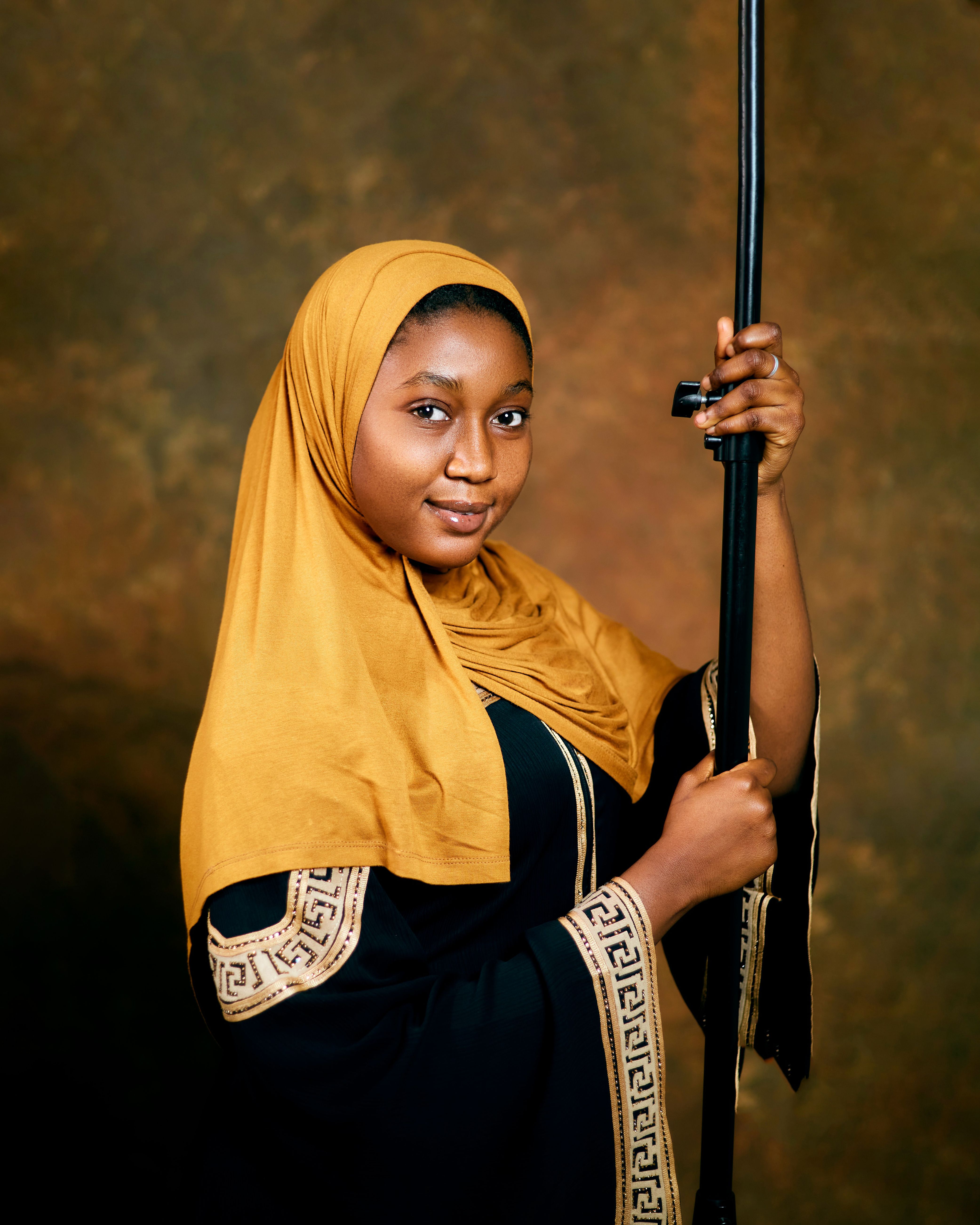 A confident woman in Nigerian fashion poses with a staff against a warm-toned background.