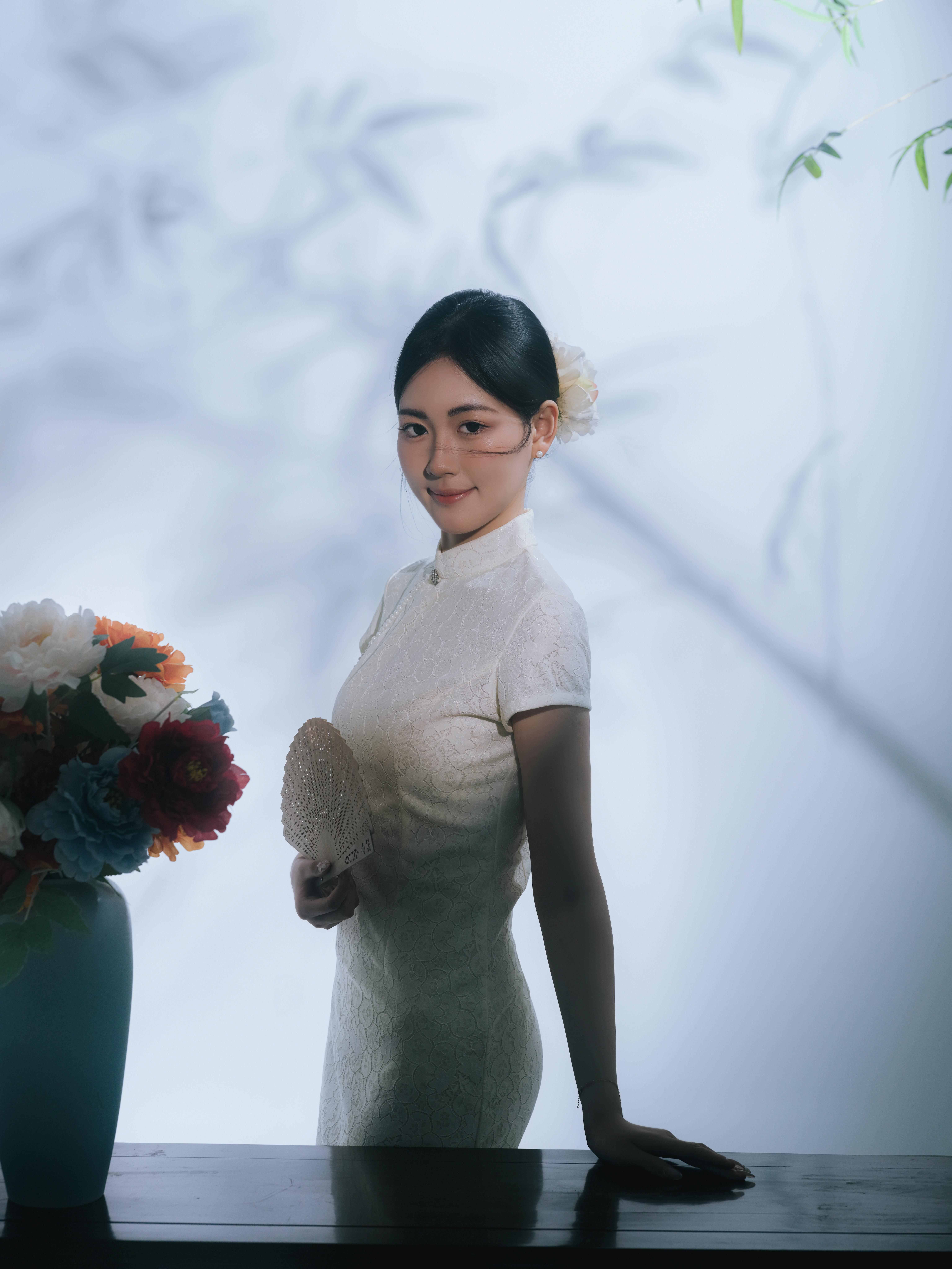 A woman in traditional dress holds a fan, standing by a vase of flowers.
