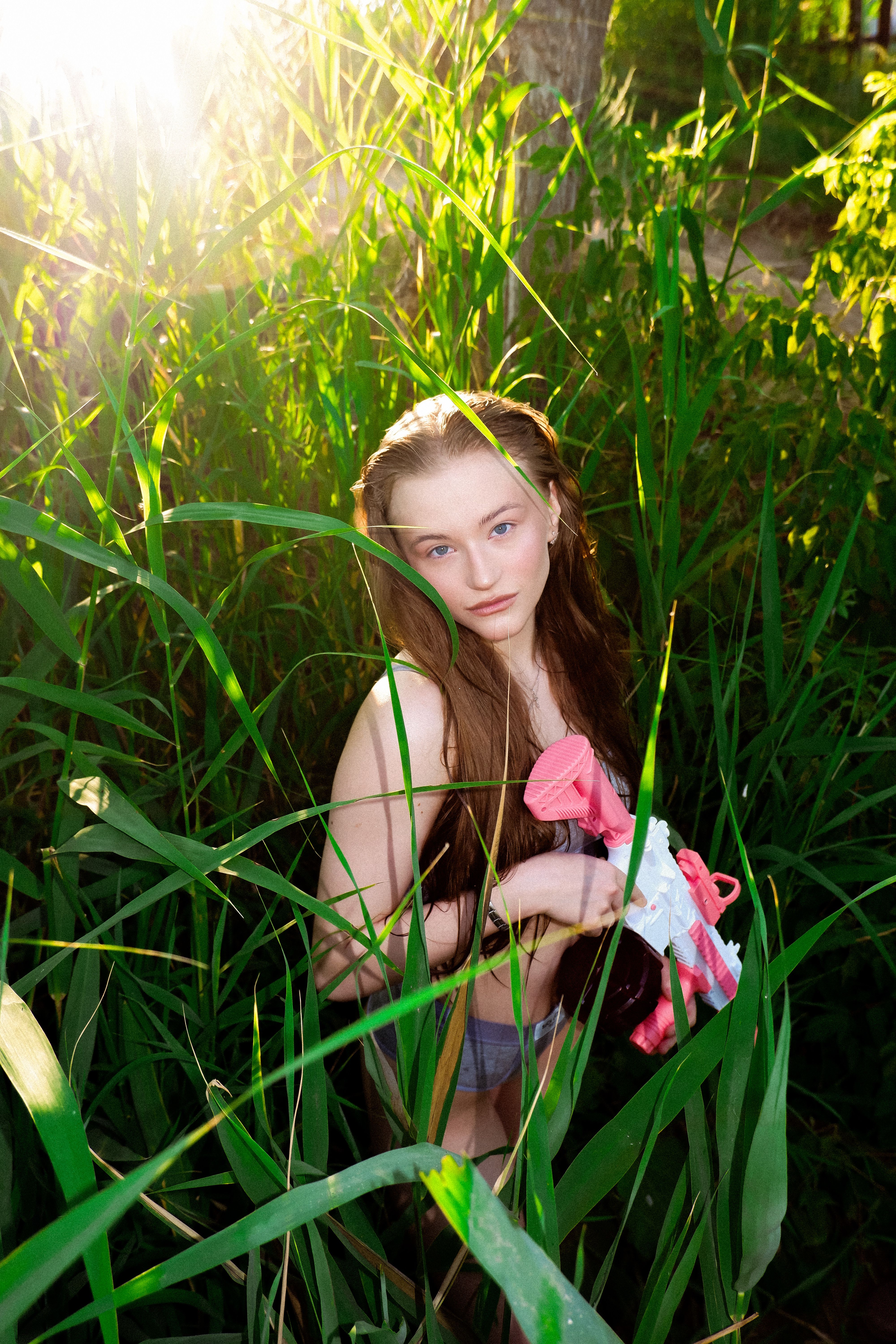 A young woman with long hair stands in lush sunlit greenery, holding a pink ribbon.