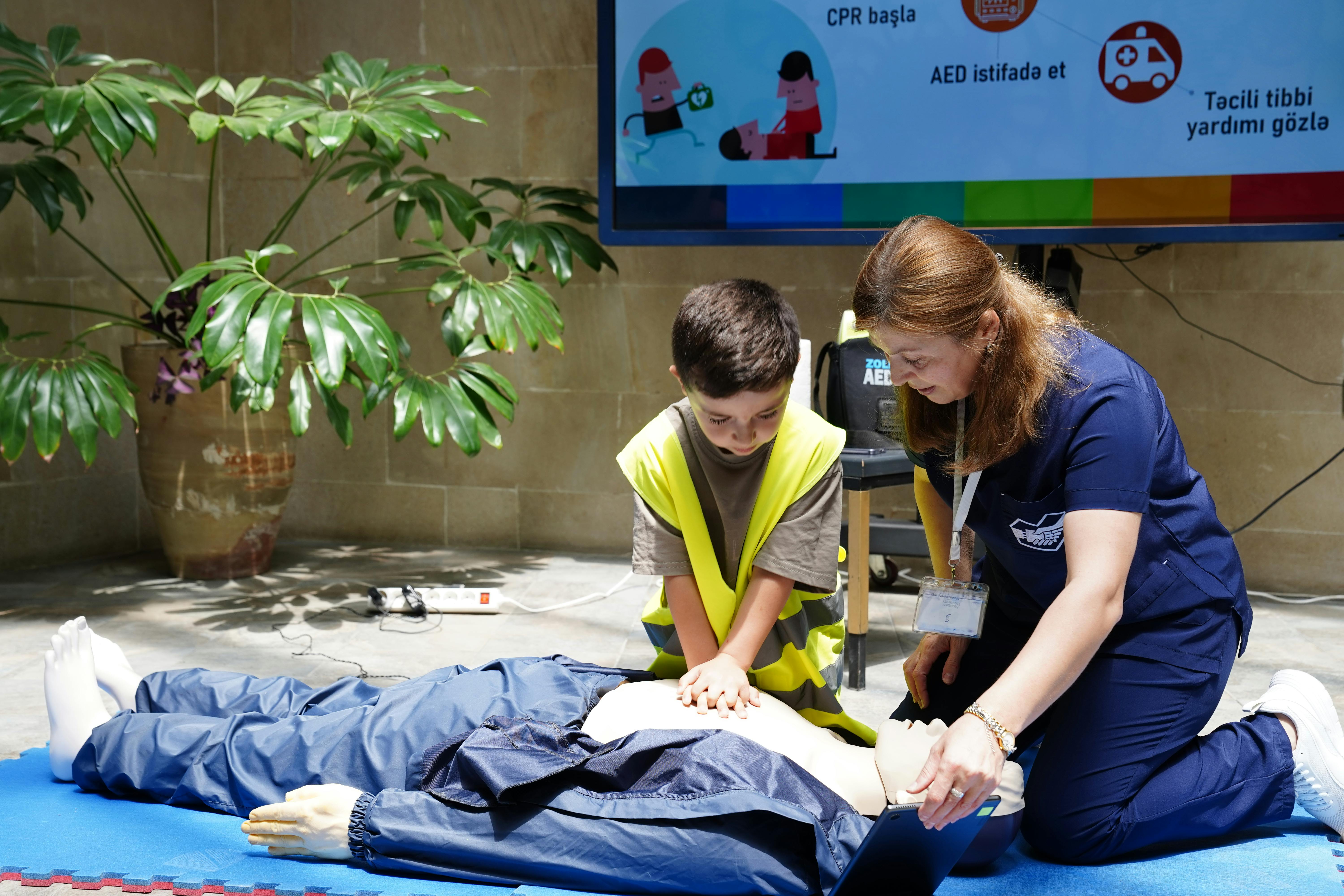 A young boy learning CPR from an instructor on a dummy indoors, with educational materials displayed.