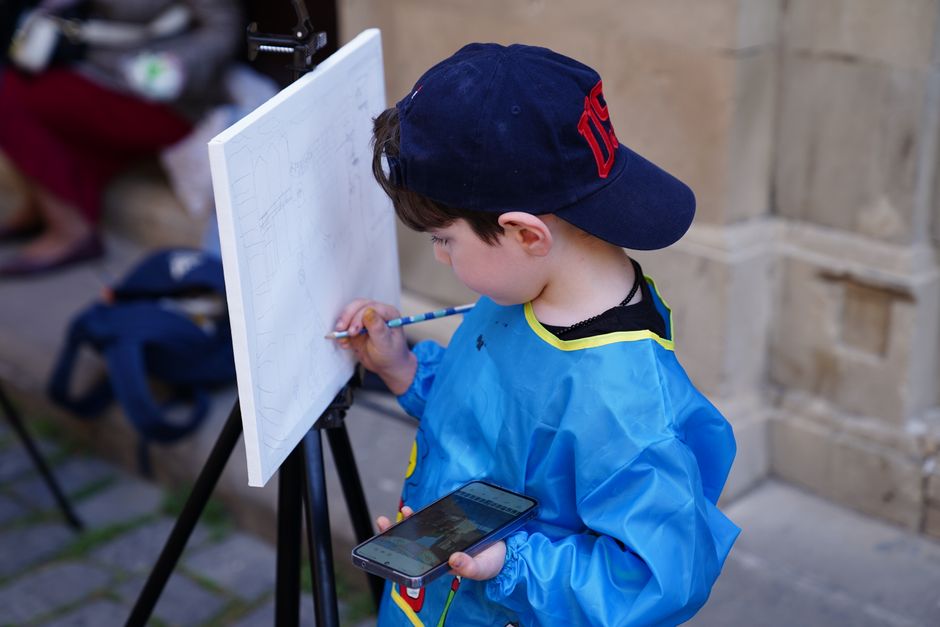 A young boy concentrates while painting outdoors, using a tablet for reference. Captured in a candid moment.