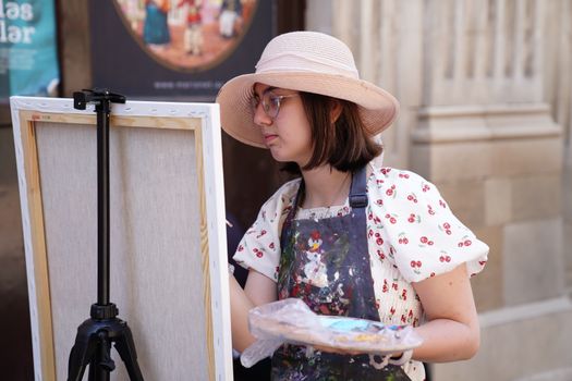 A young woman artist in a hat paints on a canvas outdoors during a sunny day.