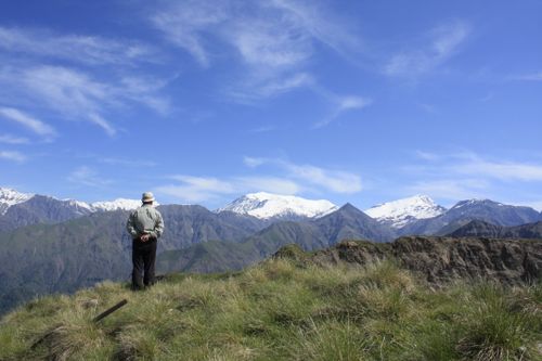 Free A man stands on a grassy hill, admiring the snow-capped mountains under a clear blue sky. Stock Photo